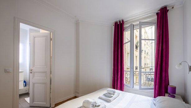Una camera d'albergo con un letto, una finestra con tende, un ingresso che porta al bagno e vista su edifici.