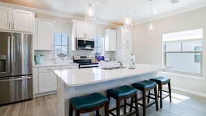 Una cocina moderna con armarios blancos, electrodomésticos de acero inoxidable, una isla grande con taburetes y luminarias colgantes.