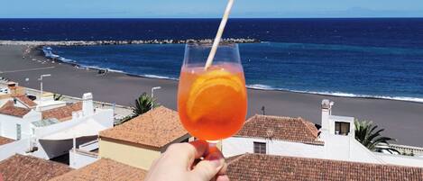 Eine Hand hält ein Glas mit einem Getränk, mit Blick auf einen Strand und Gebäude im Hintergrund.