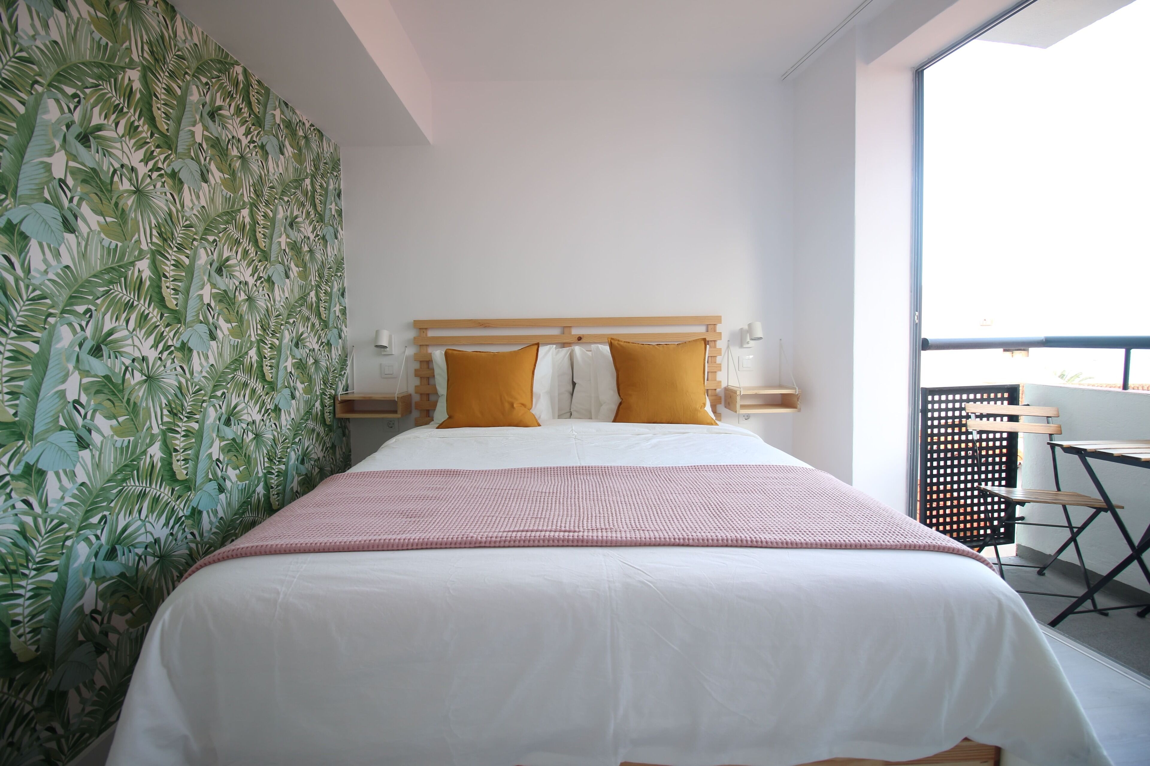 A bedroom with a large bed, two pillows, a pink blanket, a wooden headboard, and a window with a view of the outside.