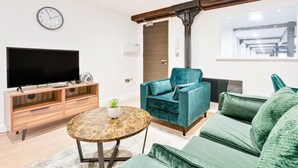 A modern living room with a flat-screen TV, wooden entertainment unit, teal and green sofas, a round coffee table, and a large window with a view of a building. - Cosy 1 Bedroom Apartment in Burton-on-Trent (Burton upon Trent)