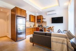 A modern kitchen with wooden cabinets, a built-in microwave, and a coffee maker. There is a gray sofa with a blanket and pillows, and a flat-screen TV mounted on the wall.