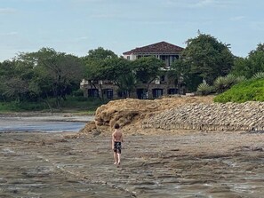 Una persona está de pie en una playa rocosa, con un edificio grande al fondo.