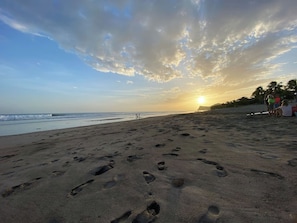 A beach with footprints leading to the water, a sunset in the background, and a few people in the distance.