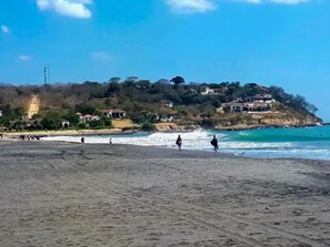 A beach with people walking, a clear sky, and houses on a hillside.