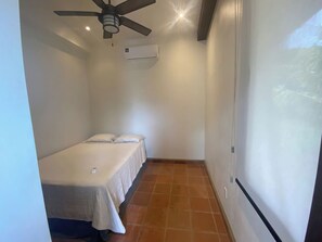Una habitación pequeña con cama individual, ventilador de techo, aire acondicionado y piso de baldosas.