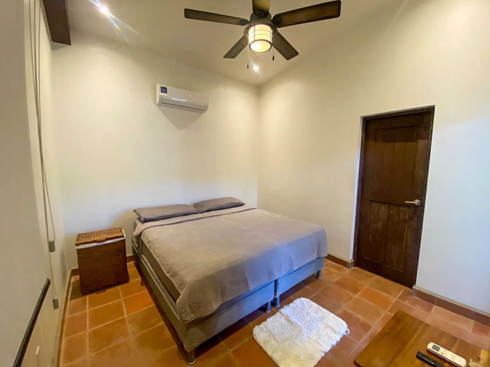 Un dormitorio con cama, ventilador de techo, equipo de aire acondicionado, puerta de madera y una mesita de noche pequeña.