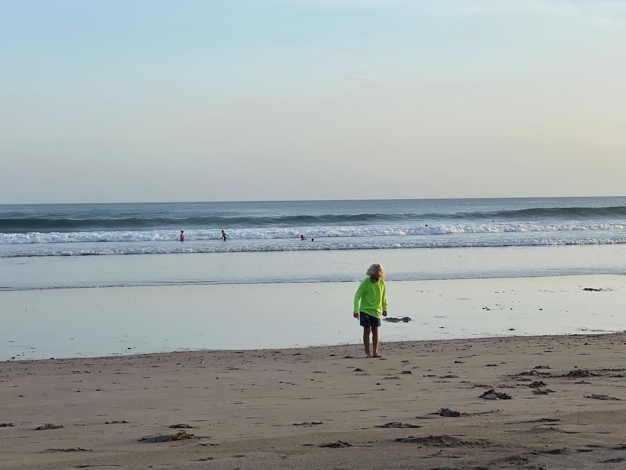 Een strand met een persoon die op het zand staat, golven op de achtergrond en een heldere lucht.