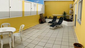 A yellow building with white shutters, a tiled patio, white plastic chairs, a small table, and two lounge chairs. - DMCharme (Ponta Delgada)