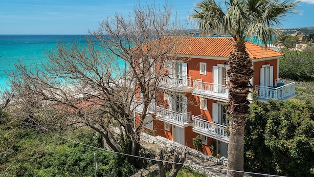 A coastal building with balconies, a palm tree, and a view of the sea. - Room “Angela Partial Sea View 8” with Sea View, Wi-Fi and Air Conditioning (kefalonia)