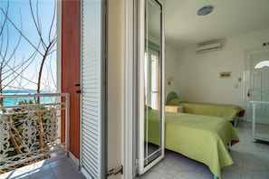 A room with a balcony, a bed with a green bedspread, a white door, and a view of the sea.