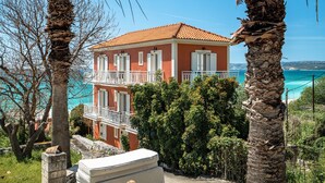 A two-story building with a balcony, surrounded by palm trees and a view of the sea. - Room “Angela partial sea view 6” with sea view, Wi-Fi, and air conditioning (kefalonia)