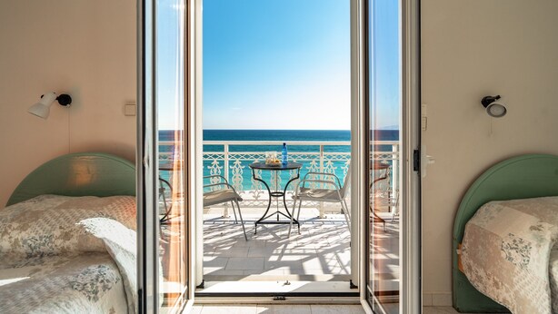 A room with a sliding glass door leading to a balcony with a table and chairs, a bed with a floral bedspread, and a view of the sea. - Room "Angela sea view 4" with sea view, Wi-Fi, and air conditioning (kefalonia)