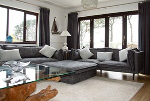 A modern living room with a sectional sofa, a glass coffee table, and a view of trees through large windows. - Hewas Water House with Pool, Hot Tub & Games Room (St Austell)