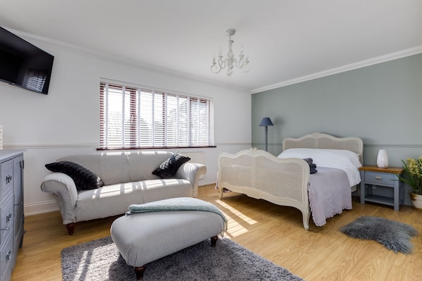 A bedroom with a white bed, a white sofa, a grey ottoman, a dresser, a TV mounted on the wall, and a chandelier. - Hewas Water House with Pool, Hot Tub & Games Room (St Austell)