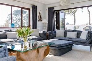 A modern living room with a glass coffee table, grey sofas, and a view of the outdoors. - Hewas Water House with Pool, Hot Tub & Games Room (St Austell)