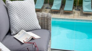 Un espace aménagé au bord de la piscine, avec un fauteuil de relaxation en osier, un coussin et une paire de lunettes posées sur l’assise.