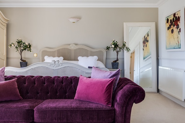A bedroom with a large bed, a purple velvet sofa, two potted plants, and a painting on the wall.