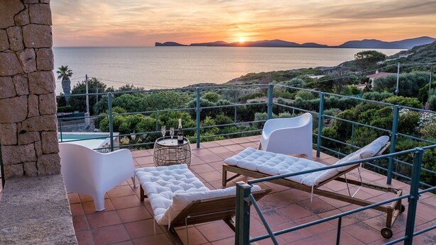 A terrace with white cushioned chairs, a small round table with a bottle and glasses, and a view of the sea at sunset. - Villa 'Coral Bay' with Sea View, Wi-Fi and Air Conditioning (Alghero)