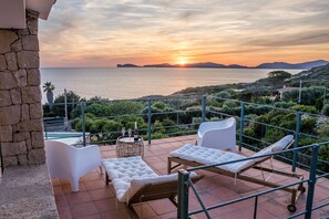 Una terrazza con sedie bianche imbottite, un tavolino rotondo con una bottiglia e dei bicchieri, e vista mare al tramonto.