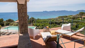 A balcony with a stone pillar, a pool, lounge chairs, and a view of the sea. - Villa 'Coral Bay' with Sea View, Wi-Fi and Air Conditioning (Alghero)