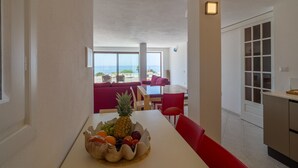 A dining area with a table set for two, a fruit bowl on the table, and a view of the ocean through large windows. - Villa 'Coral Bay' with Sea View, Wi-Fi and Air Conditioning (Alghero)