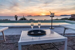 A table with two glasses of water and a teapot on a terrace overlooking a pool and sunset.