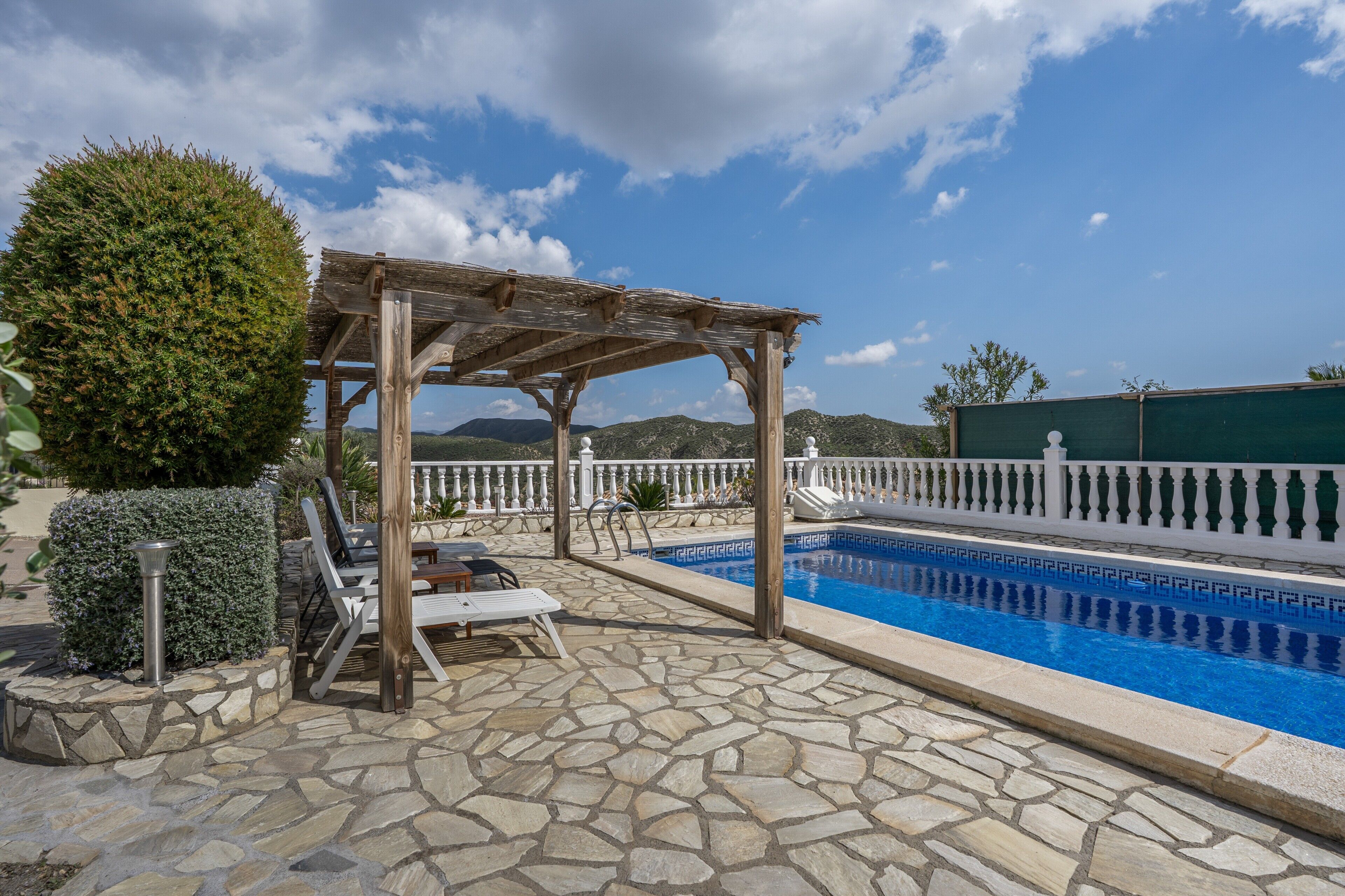 Una piscina de piedra con una pérgola y sillones de descanso, rodeada por una cerca blanca y montañas al fondo.