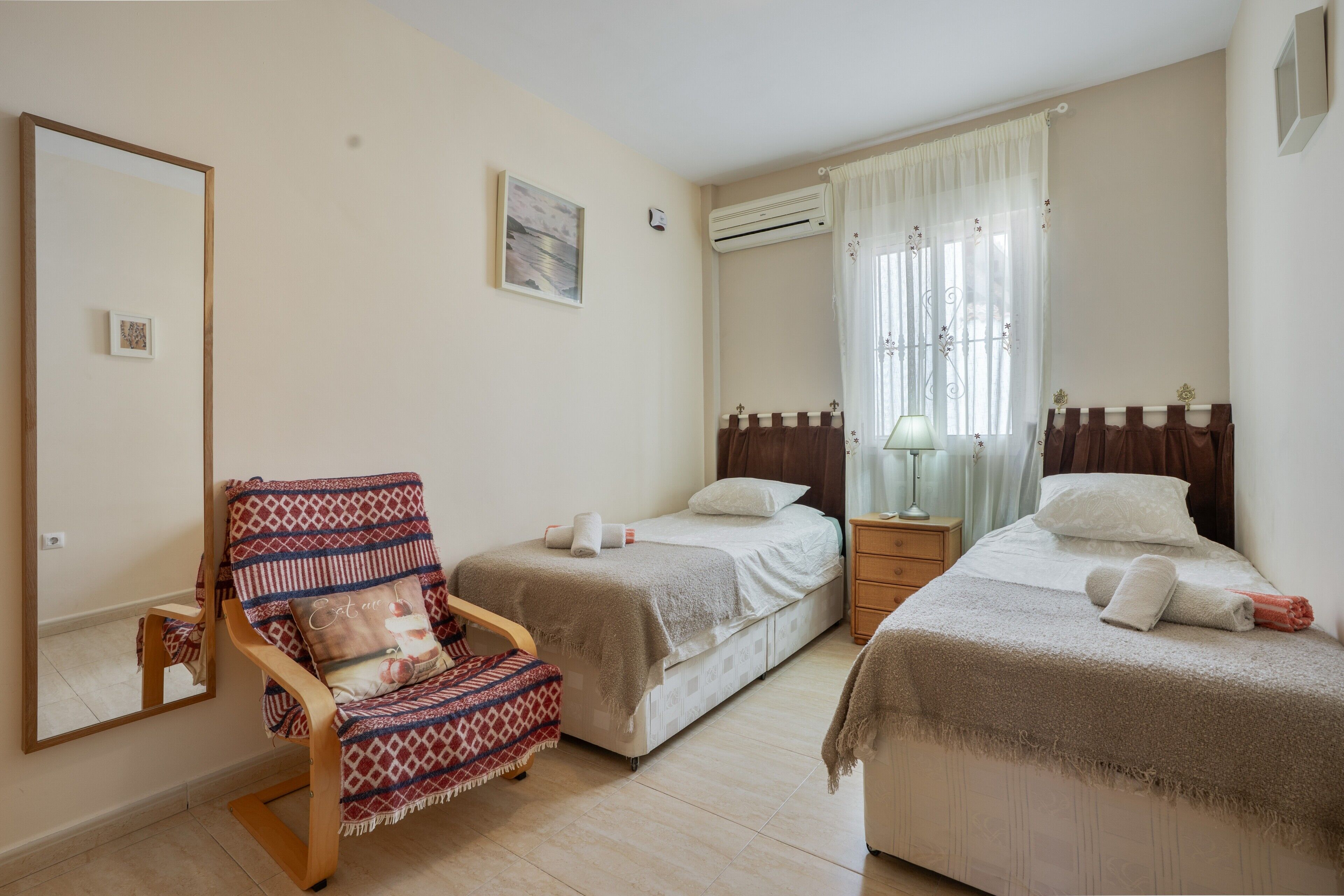 Een slaapkamer met twee bedden, een stoel, een nachtkastje, een airconditioner en een raam met gordijnen.