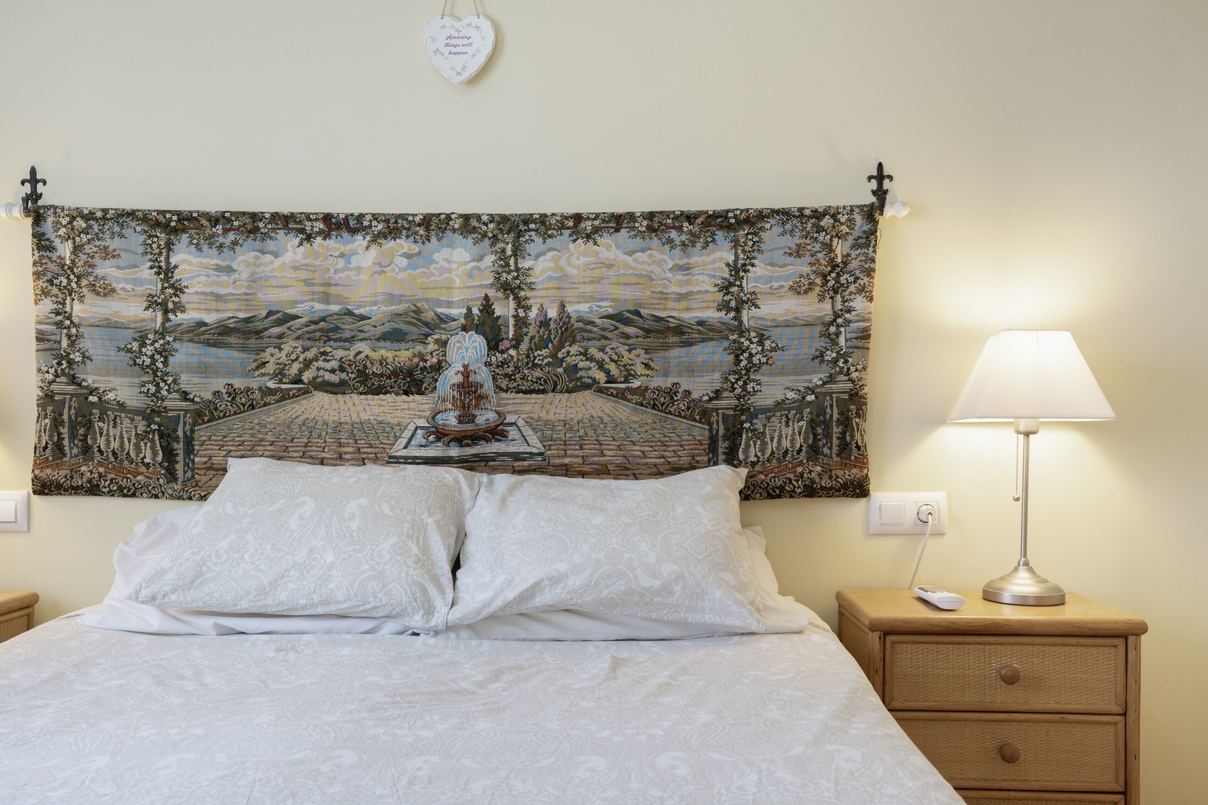 A bed with white linens, a nightstand with a lamp, and a tapestry on the wall.