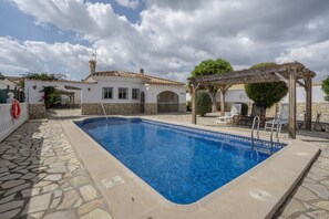 Une piscine aux bords recouverts de pierres, une pergola et un bâtiment blanc doté d’un toit en tuiles.