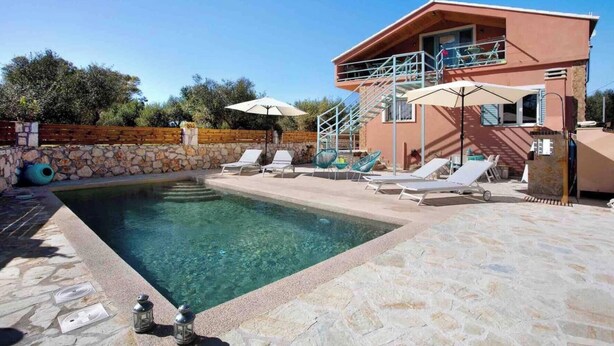 A swimming pool with lounge chairs and umbrellas, a stone wall, and a two-story building with a balcony. - Zante Loft Amici with Pool, Garden, Terrace and Wi-Fi (Zakynthos)
