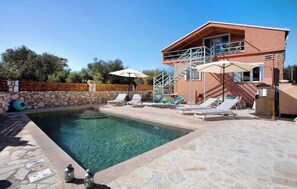 En swimmingpool med liggestole og parasoller, en stenvæg og en toetagers bygning med balkon.