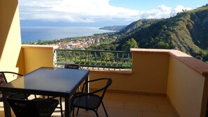 A balcony with a table and chairs, overlooking a coastal town and hills.
