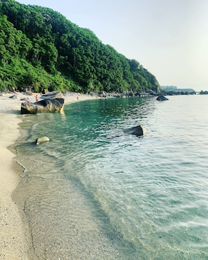 A beach with clear water, rocks, and a forested hillside.