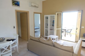 A living room with a beige sofa, a dining table with chairs, a bookshelf, and a sliding glass door leading to an outdoor area.