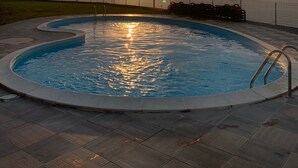 A swimming pool with a view of the sunset over the ocean, surrounded by a fence and a grassy area.
