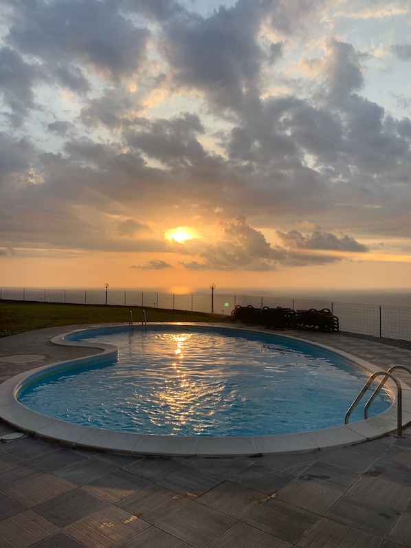 A swimming pool with a view of the sunset over the ocean, surrounded by a fence and a grassy area.