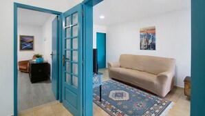 An open door leading to a living room with a beige sofa, a patterned rug, and a television on a stand. - Country House 'Tibagota' with Mountain View, Wi-Fi and Air Conditioning (São Brás de Alportel)
