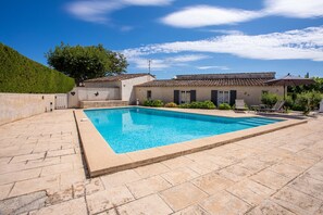 A rectangular swimming pool with a tiled deck, a building with a tiled roof, and a green hill in the background.