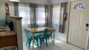 A dining area with a wooden table and four teal chairs, a small kitchen area with a stove, and a white door with a stained glass window. - 08) The Marlin Cottage (Inglis)