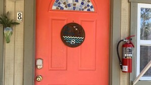 A bright red door with a mosaic window above it, a round sign with a wave design, and a fire extinguisher mounted on the wall to the right. - 08) The Marlin Cottage (Inglis)