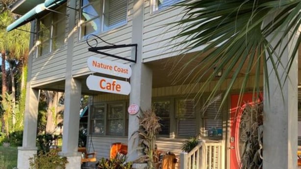 A building with a sign reading "Nature Coast Cafe" and a porch with potted plants and pumpkins.