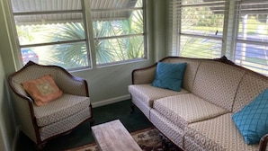 A living room with a patterned sofa and armchair, a wooden coffee table, and a view through large windows.