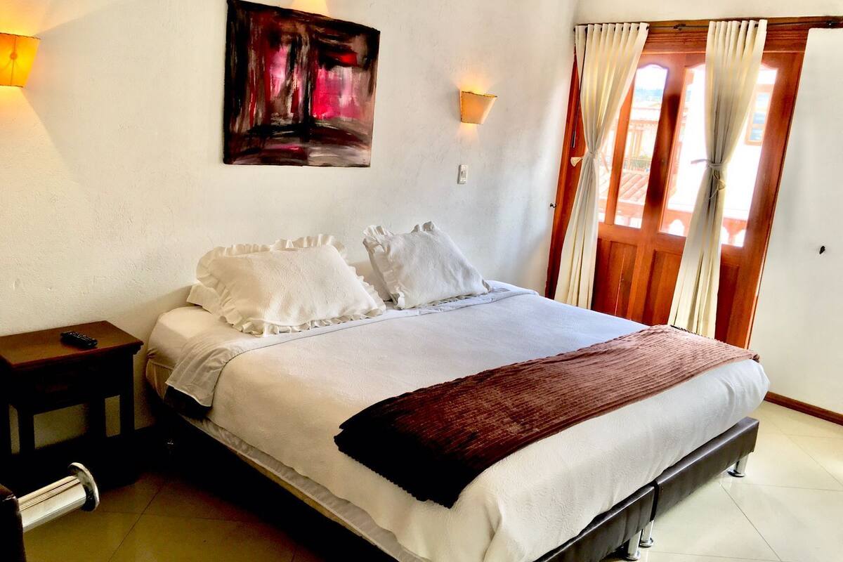 Una cama bien hecha con ropa blanca y un plaid marrón, una mesita de noche con un control remoto, y un cuadro en la pared.