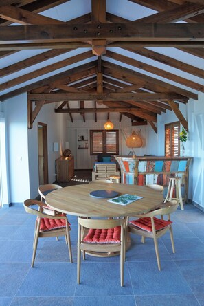 A dining area with a wooden table and chairs, a high ceiling with exposed beams, and a view of the adjacent room.