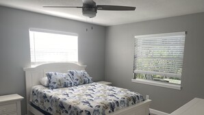 Un dormitorio con una cama, mesitas de noche, ventilador de techo y dos ventanas con persianas.
