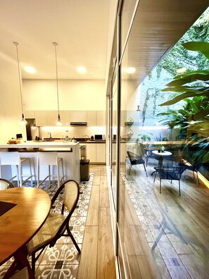 A modern kitchen with white cabinets, a central island, pendant lighting, and a view of a lush garden through large glass doors.