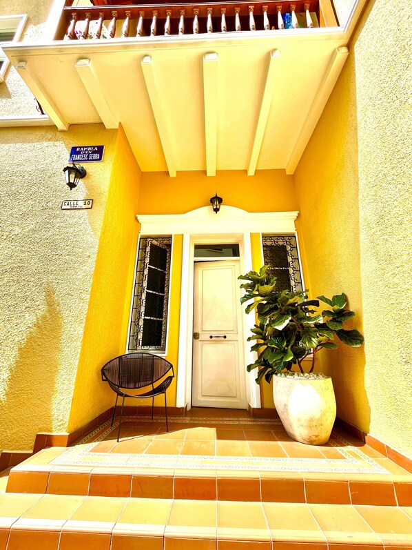 A yellow building with a white door, a potted plant, and a chair outside.