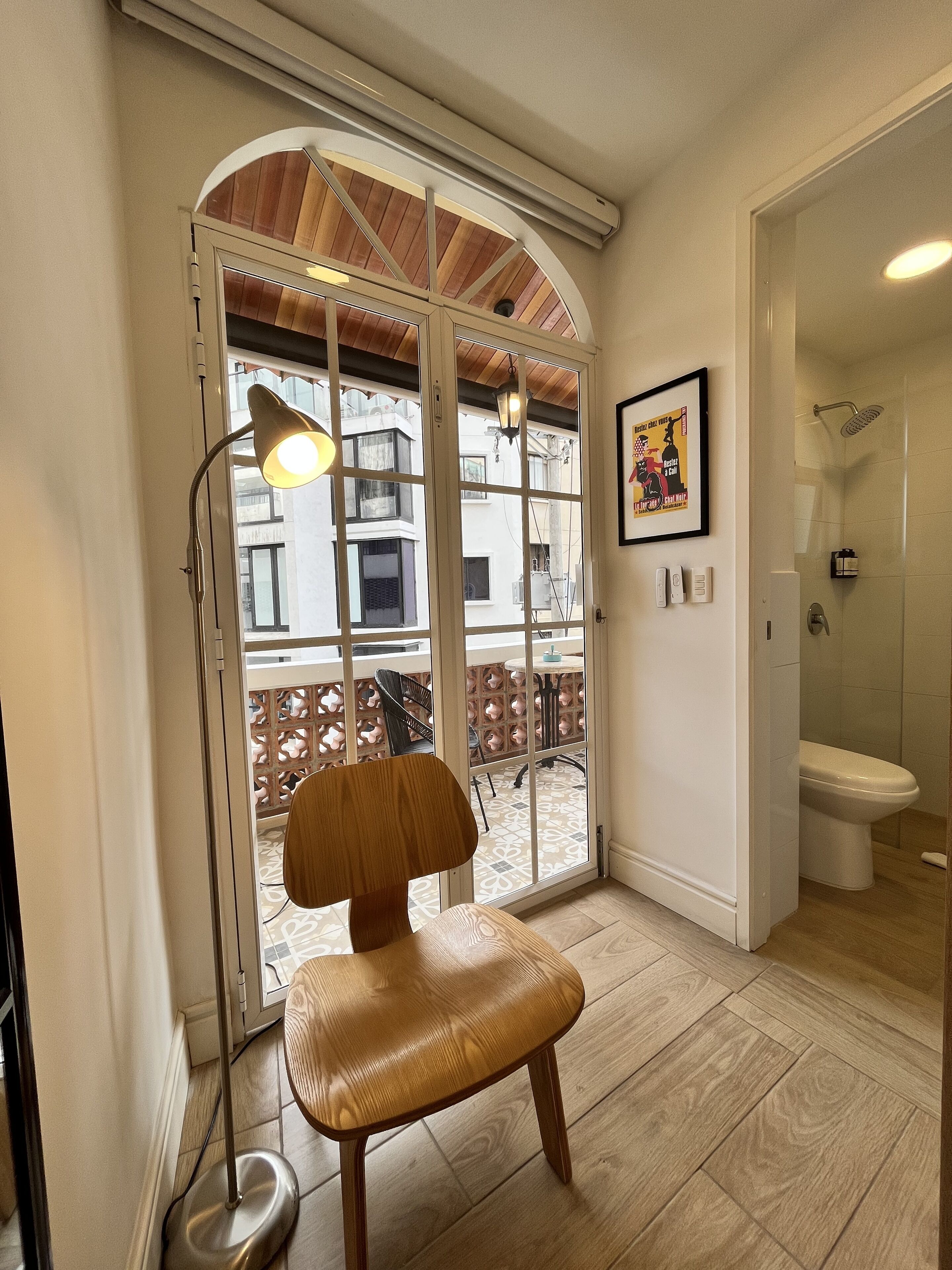 A room with a wooden chair, a floor lamp, a large window, and a view of the outside balcony.